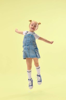 Kot elbiseli mutlu kız çocuğu sevinçle atlıyor, sarı arka planda eğlence ve mutluluğu ifade ediyor. Çocukluk kavramı, yaz tatili, moda, eğlence, eğlence