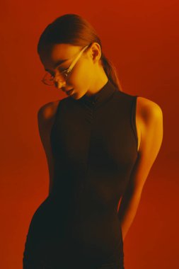 Atkuyruklu, şeffaf gözlüklü ve kolsuz mayo giyen, neon ışıkta kırmızı arka planda poz veren çekici bir kadının portresi. Moda, tarz, gençlik kültürü kavramı