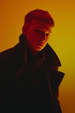 Siyah ceketli bir gencin portresi, kameraya bakıyor, sarı stüdyonun arka planında neon ışıkta poz veriyor. Cesur moda kavramı, tarz, gençlik kültürü, kendini ifade etme