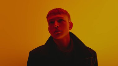 Siyah ceketli ve kısa saçlı şık bir gencin portresi, yukarı bakıyor, neon ışıkta sarı arka plana karşı poz veriyor. Cesur moda, tarz, gençlik kültürü, kendini ifade etme kavramı