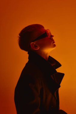 Kısa saçlı, siyah ceketli ve fütüristik büyük güneş gözlüklü genç adamın portresi turuncu stüdyo arka planına bakıyor. Cesur moda, tarz, gençlik kültürü, kendini ifade etme