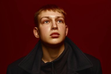 Koyu renk ceketli genç adamın yakın plan portresi zengin kırmızı arka planda poz veriyor, yukarı doğru yoğun bir ifadeyle bakıyor. Modern moda kavramı, gençlik kültürü, modern tarz.