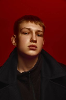 Siyah ceketli, koyu kırmızı arka planda duran ve doğrudan kameraya bakan kendine güvenen genç adamın yakın portresi. Cesur moda kavramı, tarz, gençlik kültürü, kendini ifade etme
