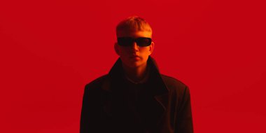 Siyah ceketli ve güneş gözlüklü genç bir adamın portresi kasvetli kırmızı arka planda ileri bakıyor. Güzellik, moda, gençlik kültürü, fütüristik atmosfer, görsel kimlik, modern eğilimler kavramı