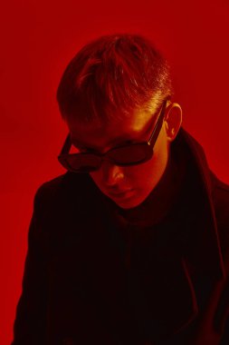 Güneş gözlüğü takan ve siyah ceketli genç adam, kırmızı stüdyo arka planına karşı koyu kırmızı ışık altında poz veriyor. Cesur moda kavramı, tarz, gençlik kültürü, kendini ifade etme