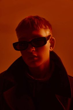 Modayı takip eden güneş gözlüklü ve siyah ceketli genç bir adamın yakın plan portresi, neon ışıkta kırmızı stüdyoda poz veriyor. Cesur moda kavramı, tarz, gençlik kültürü, kendini ifade etme