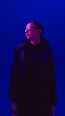 Güneş gözlüğü takan ve siyah ceketli genç bir kadının portresi neon ışıkta mavi arka planda poz veriyor. Cesur moda kavramı, tarz, gençlik kültürü, kendini ifade etme