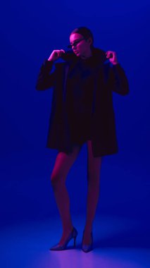 Siyah ceketli, güneş gözlüklü ve topuklu ayakkabılı, neon ışıkta mavi arka planda poz veren zarif genç bir kadının tam boy portresi. Cesur moda anlayışı, tarz, gençlik kültürü, kendini ifade etme, güzellik