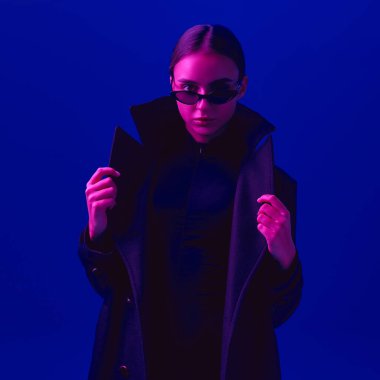 Siyah ceketli, güneş gözlüklü, kendine güvenen genç bir kadının portresi neon ışıkta mavi arka plana bakıyor. Cesur moda anlayışı, tarz, gençlik kültürü, kendini ifade etme, güzellik