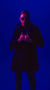 Siyah ceketli ve güneş gözlüklü genç bir kadının portresi neon ışıkta mavi arka planda kendine güvenen bir şekilde poz veriyor. Cesur moda anlayışı, tarz, gençlik kültürü, kendini ifade etme, güzellik