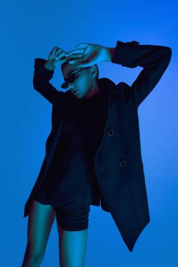 Kendine güveni tam genç bir kadın, ceketli ve güneş gözlüklü mavi arka planda, neon ışıkta dinamik bir pozda duruyor. Cesur moda anlayışı, tarz, gençlik kültürü, kendini ifade etme, güzellik