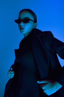 Güneş gözlüklü, siyah ceketli ve takım elbiseli, mavi arka planda neon ışıkta poz veren kendinden emin genç bir kadının portresi. Cesur moda anlayışı, tarz, gençlik kültürü, kendini ifade etme, güzellik