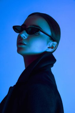 Güneş gözlüklü ve siyah ceketli zarif bir genç kadının yakın plan portresi neon ışıkta mavi arka planda güvenle poz veriyor. Cesur moda kavramı, gençlik kültürü, kendini ifade etme, güzellik