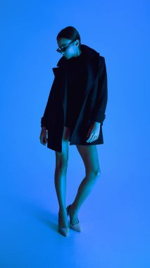 Paltolu, topuklu ve güneş gözlüklü kendine güvenen genç bir kadının mavi arka planda durduğu uzun bir portre. Cesur moda anlayışı, tarz, gençlik kültürü, kendini ifade etme, güzellik