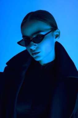 Güneş gözlüklü ve siyah ceketli zarif genç kadının yakın plan portresi neon ışıkta mavi arka planda güvenle poz veriyor. Cesur moda kavramı, gençlik kültürü, kendini ifade etme, güzellik