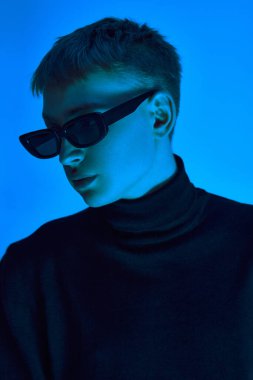 Güneş gözlüklü ve siyah ceketli şık bir gencin yakın plan portresi neon ışıkta mavi arka planda güvenle poz veriyor. Cesur moda kavramı, gençlik kültürü, kendini ifade etme, güzellik