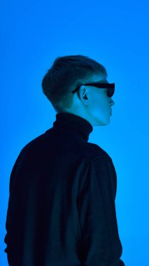 Balıkçı yaka ve güneş gözlüklü şık bir gencin yan profili, derin neon mavisi arka plan altında poz vermesi fütüristik zarafeti ifade ediyor. Cesur moda kavramı, tarz, gençlik kültürü, kendini ifade etme