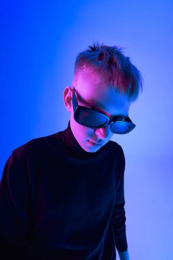 Kısa saçlı, güneş gözlüğü takan ve balıkçı yaka kazak giyen neon ışıkta mavi arka planda poz veren şık bir gencin yakın plan portresi. Cesur moda, tarz, gençlik kültürü kavramı