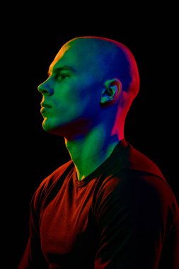 Siyah tişörtlü yakışıklı kel bir adamın karanlık arka planda kendine güvenen ve neon ışıkta ciddi bir ifadeyle poz verişinin yan görüntüsü. Erkek güzelliği, moda, tarz, gençlik kültürü kavramı