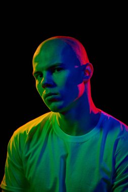 Beyaz tişörtlü genç kel adamın ön açılı portresi, çok renkli neon ışıkta, karanlık arka planda nötr bir ifadeyle kameraya doğru bakıyor. Erkek güzelliği, moda, tarz, gençlik kavramı