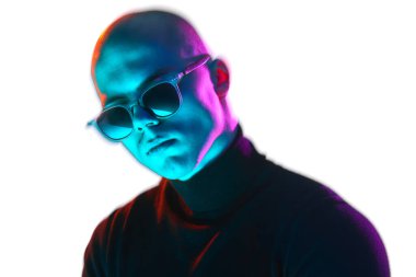 Siyah balıkçı yaka yaka ve güneş gözlüklü yakışıklı genç bir adam portresi, mavi ve pembe neon ışıkta beyaz arka planda güvenle poz veriyor. Erkek güzelliği, moda, tarz, gençlik kültürü kavramı