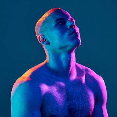 Kaslı vücutlu yakışıklı, kel bir adamın portresi, üstsüz poz veriyor, mavi arka planda nötr bir ifadeyle neon ışıkta yukarı bakıyor. Erkek güzelliği, moda, tarz, gençlik kültürü kavramı
