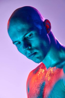 Üstsüz poz veren, kendine güvenen kel bir gencin yakın plan portresi, neon ışıkta açık mor arka planda ciddi bir ifadeyle aşağıya bakıyor. Erkek güzelliği, moda, tarz, gençlik kültürü kavramı