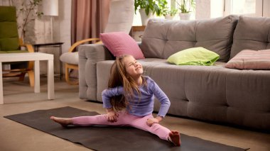 Evde, oturma odasında yoga minderinin üzerinde mutlu bir ifadeyle başını kaldıran küçük bir çocuk. Fiziksel aktivite, egzersiz, esneklik ve sağlıklı, aktif yaşam tarzı. Çocukluk kavramı