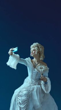 Klasik elbise ve beyaz peruklu asil bayan mavi arka planda telefon ve ayna eşliğinde selfie çekiyor. Sosyal medya paylaşımı, blog yazarı. Dönem, güzellik, tarih, sanat, yaratıcılık, ağ karşılaştırması