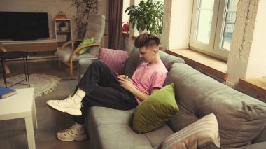 Pembe gömlekli bir adam evdeki rahat oturma odasındaki modern kanepede uzanıyor, akıllı telefonlu, konsantre görünüyor. Teknoloji kavramı, dijital yaşam tarzı, gençlik, iletişim, mobil kullanım