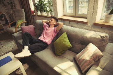 Elleri başının arkasında, kulaklık takmış, rahat bir oturma odasında dinlenen bir adam. Dinlenme konsepti, sakinlik, müzik dinleme, dijital eğlence, kapalı yaşam tarzı, zihinsel şarj.