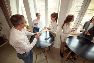 Bir grup iş arkadaşı modern ofiste yüksek masaların etrafında durur, teneffüste konuşur ve kahve içerler. Ağ kurma, kırma, gayri resmi toplantı, ofis hayatı, gündelik tartışma kavramı.