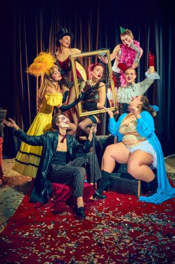 Dramatik klasik giysiler içindeki erkek ve kadınlar, kırmızı halı üzerindeki altın çerçeveyle etkileşimli bir şekilde poz veriyorlar. Konfeti ile kaplanmış, klasik performansın tiyatral duyguları. Kabare, gösteri, sirk, retro konsepti