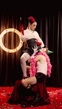 İki kabare sanatçısı, erkek ve kadın, grevdeki vintage kostümler içinde, konfeti kaplı kırmızı halıda dramatik bir poz veriyorlar. Kabare, gösteri, sirk, retro, vintage, performans konsepti.