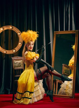 Sarı, klasik tarzda giyinmiş, kırmızı halıdaki sandalyeye zarif bir şekilde oturmuş, uzun sigara tutacağıyla poz veren zarif bir kadın sanatçı. Kabare, gösteri, sirk, retro, performans kavramı.