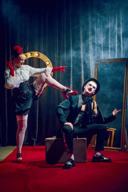 Kabaredeki kadın, kırmızı eldivenli, bavullu, dumanlı ışıklarla oturan adama doğru bacaklarını kaldırıyor. Kabare, tiyatro, mizah, bürlesk, gösteri, kırmızı halı, tarz, makyaj, ifade.