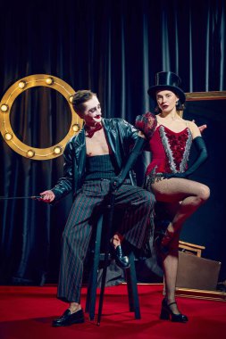 Erkek ve kadın kabare sanatçısı, silindir şapka ve parlak kırmızı elbise de dahil olmak üzere çarpıcı bir klasik giysi giyerek kırmızı halıda dinamik bir şekilde poz veriyor. Gösteri konsepti, sirk, retro, vintage, performans.