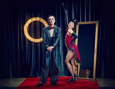 Tarz sahibi kabare çifti ışıkların altında teatral kıyafetler kamış şapkası ve dramatik ifadeler takınarak poz veriyor. Kabare, gösteri, tiyatro, vintage, retro, performans, mizah, karakter, ifade.