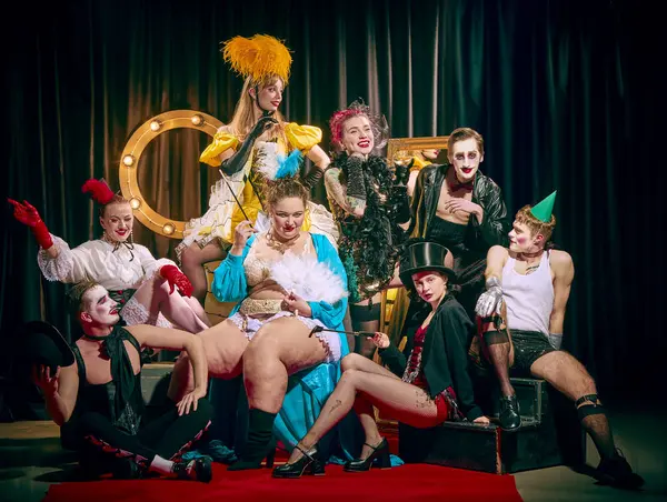 Özenle hazırlanmış klasik kostümlü büyük bir kabare grubu kırmızı halıda birlikte poz vererek tiyatral ifadeler sergiliyor. Kabare, gösteri, sirk, retro, vintage, performans konsepti.
