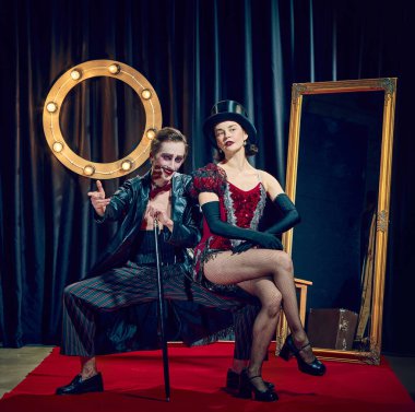 Oturan kabare göstericileri, şık giyinen kadın ve esmer retro tiyatro geçmişine karşı baston pozu veren çarpıcı makyajlı adam. Gösteri konsepti, sirk, retro, vintage, performans.