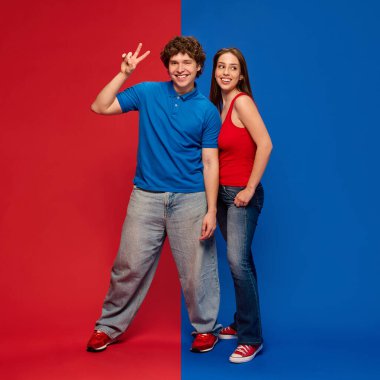 Mutlu gençlerin tam boy portresi, barış işareti gösteren bir adam, cesur kırmızı mavi arka plana karşı gülümseyen bir kadın, neşe ve gençlik. İnsan duyguları kavramı, ilişki, yaşam tarzı, moda