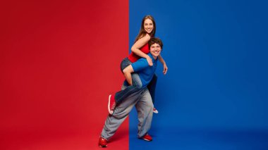 Gülen kadın, erkeklerin sırtına biniyor. Kırmızı mavi arka planda enerji ve neşeyle poz veriyor. Sıradan giysili bir çift. İnsan duyguları kavramı, ilişki, yaşam tarzı, moda, eğlence, oyunculuk