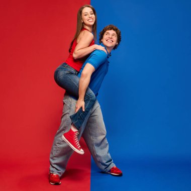 Genç bir adamın genç bir kadını sırtında taşıması. Hem yukarı bakıyor, hem gülümsüyor, canlı kırmızı ve mavi arka plana karşı. İnsan duyguları kavramı, ilişki, yaşam tarzı, moda, eğlence