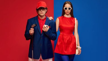 Golf sopası tutan şık bir adam ve kırmızı arka planda kendine güvenen güneş gözlüğü takan bir kadın. Spor, hobi, insan duyguları, ilişki, yaşam tarzı, moda kavramı