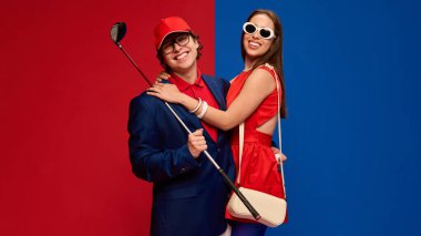 Mutlu çift sarılıyor, ellerinde golf sopasıyla gülümsüyor, kırmızı mavi arka planda şık kıyafetler giyiyorlar. İnsan duyguları kavramı, ilişki, yaşam tarzı, moda, spor, eğlence
