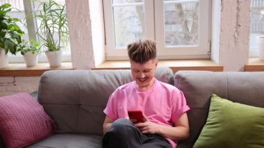 Pembe tişörtlü genç adam rahat gri koltukta oturuyor, elinde akıllı telefon, gülümsüyor, neşeli bir ruh hali var. Dijital yaşam tarzı, konfor, bağlantı, iletişim, olumlu duygu, boş zaman