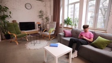 Pembe gömlekli bir adam evdeki rahat oturma odasındaki modern kanepede uzanıyor, akıllı telefonlu, konsantre görünüyor. Teknoloji kavramı, dijital yaşam tarzı, gençlik, iletişim, mobil kullanım