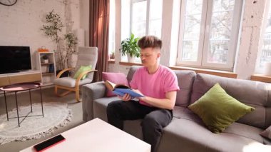 Pembe tişörtlü genç adam gri kanepede oturur, güneşli oturma odasında kitap okur. Eğitim kavramı, rahatlama, evde öğrenim, dijital detoks, bilgi, ev içi rahatlık, konfor.