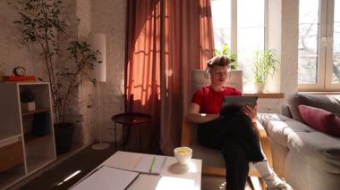 Kırmızı tişörtlü genç adam sandalyede oturuyor, kulaklık takıyor, güneş ışığı odasında tablet kullanıyor. Kahve fincanı ve defter masanın üstünde. Uzaktan çalışma kavramı, çevrimiçi öğrenme, dijital yaşam tarzı, teknoloji