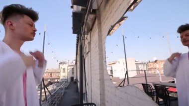 Çatıda beyaz önlüklü genç adam tasmayı ayarlıyor, duvardaki aynaya bakıyor. Arka planda binalar ve sicim ışıkları var. Tarz, güven, kentsel yaşam tarzı, özbakım, moda kavramı.